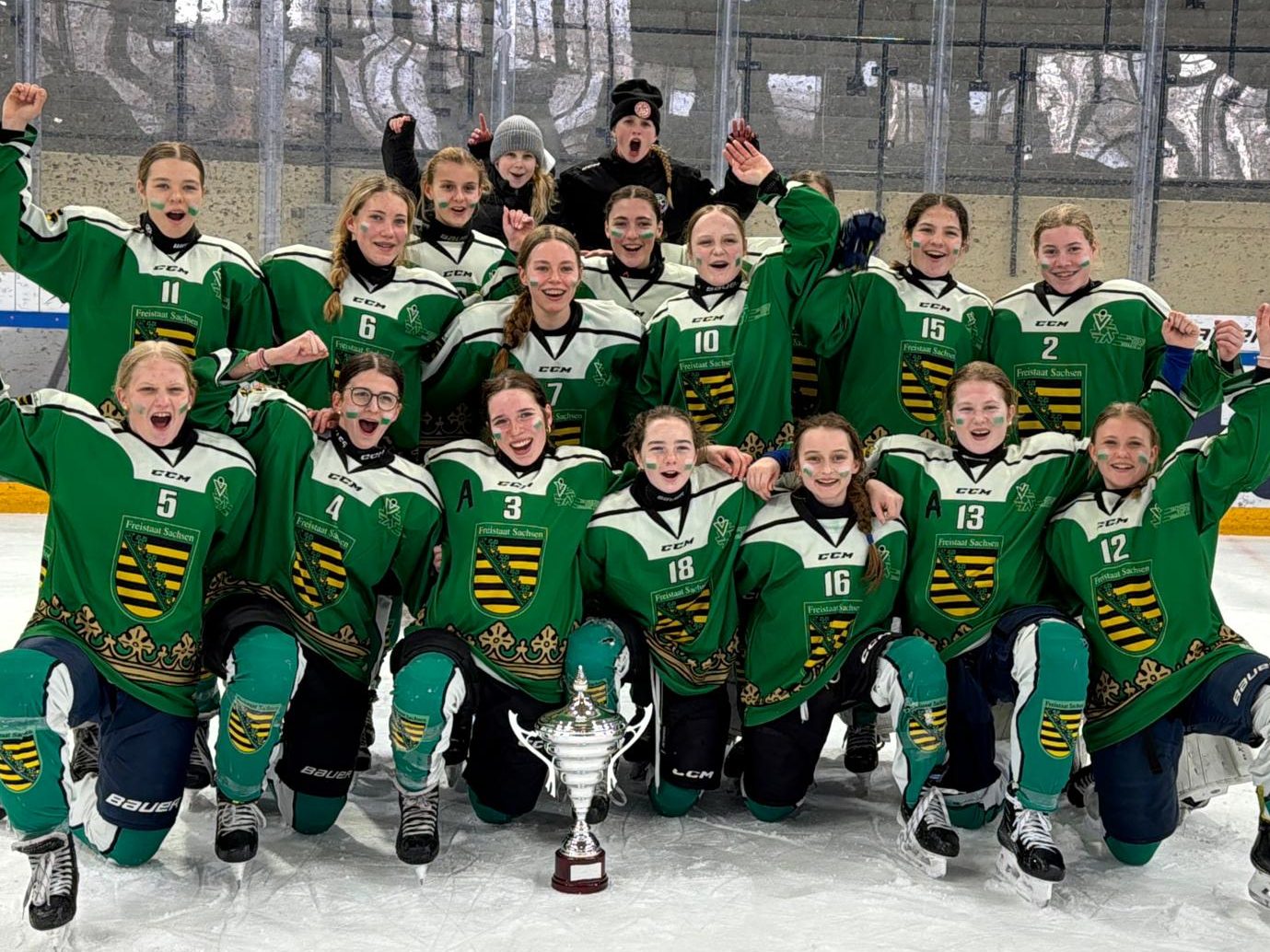 Gruppenbild der freudestrahlenden Eishockey-Nachwuchsauswahl Sachsen/Berlin mit Pokal nach dem Sieg beim Länderpokal in Füssen. Das Team in grünen Trikots jubelt mit hoch erhobenen Armen vor der Bande.